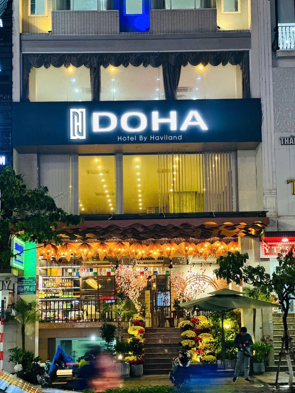The hotel's exterior features a modern façade with the name 'DOHA' prominently displayed above the entrance. Warm lighting and decorative plants enhance the welcoming atmosphere. A staircase leads to the entrance, flanked by vibrant flowers and outdoor seating under umbrellas.