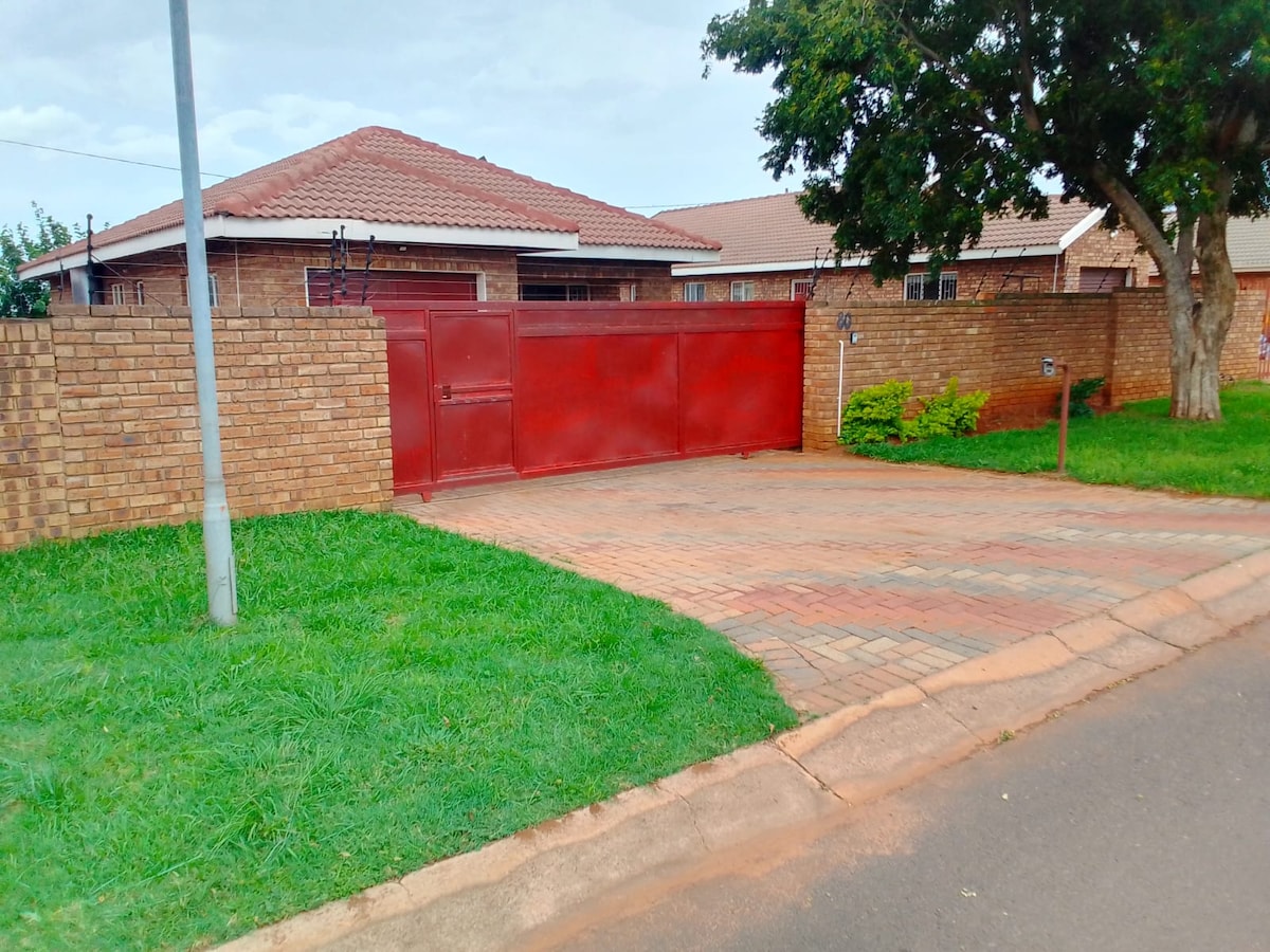 A well-maintained exterior features a red gate set into a brick wall, providing security and privacy. A paved driveway leads up to the entrance, framed by neatly trimmed grass. Surrounding homes are visible, showcasing a residential neighborhood.