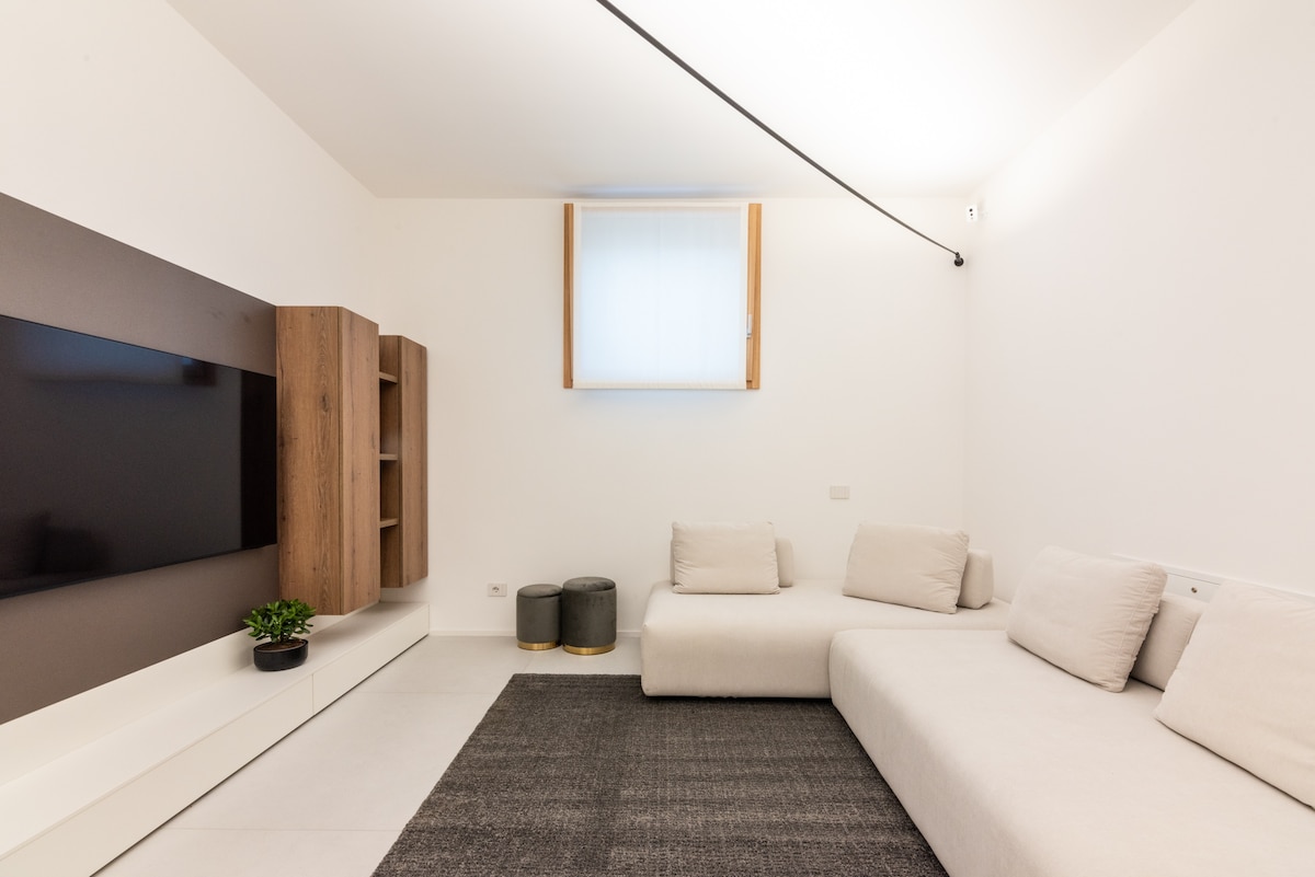 A minimalist living area features a large, wall-mounted television opposite a spacious sectional sofa with soft cushions. A plush rug adds texture to the floor, while a light, airy window allows natural light to fill the room.
