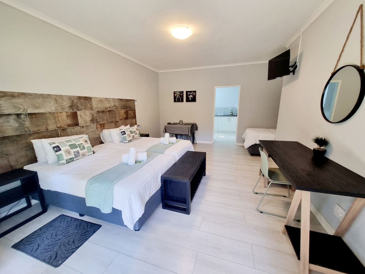 A spacious room features three 3/4 beds with neutral bedding, complemented by a wooden accent wall. An area for dining or work includes a table and chairs. A kitchenette is visible in the background, while a smart TV is mounted on the wall.