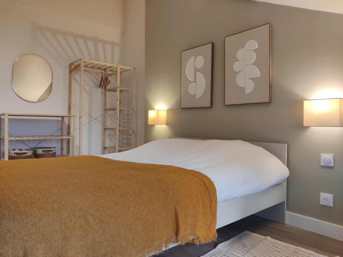 A softly lit bedroom features a spacious bed covered with a white duvet and an orange throw blanket. Elegant artworks are displayed above the bed, complemented by two bedside lamps. A clothing rack and shelving unit provide practical storage, enhancing the room's functionality.