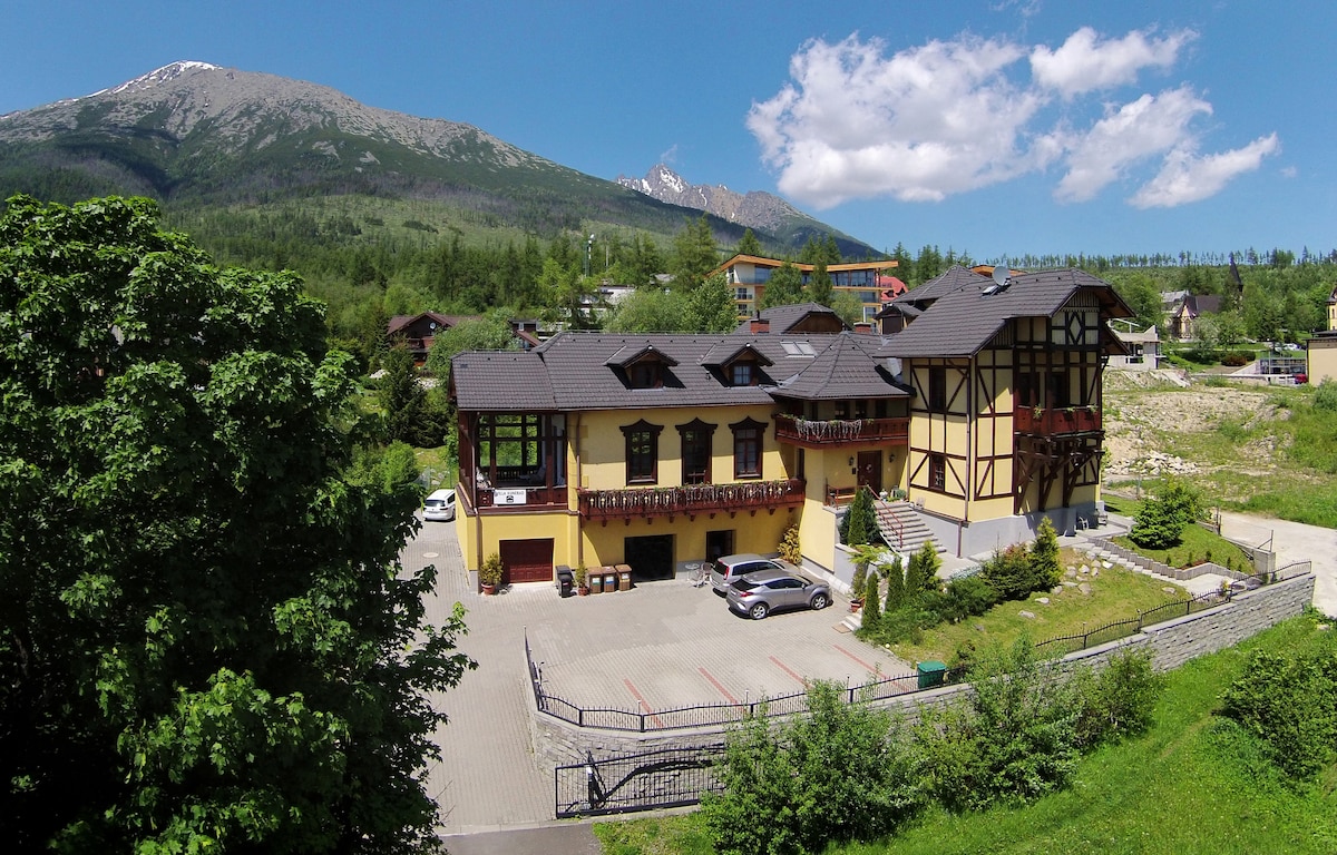 The exterior of the villa is characterized by a combination of colorful traditional architecture and modern design elements. Surrounding greenery complements the parking area in front, which accommodates multiple vehicles. Majestic mountains provide a scenic backdrop under a clear blue sky.