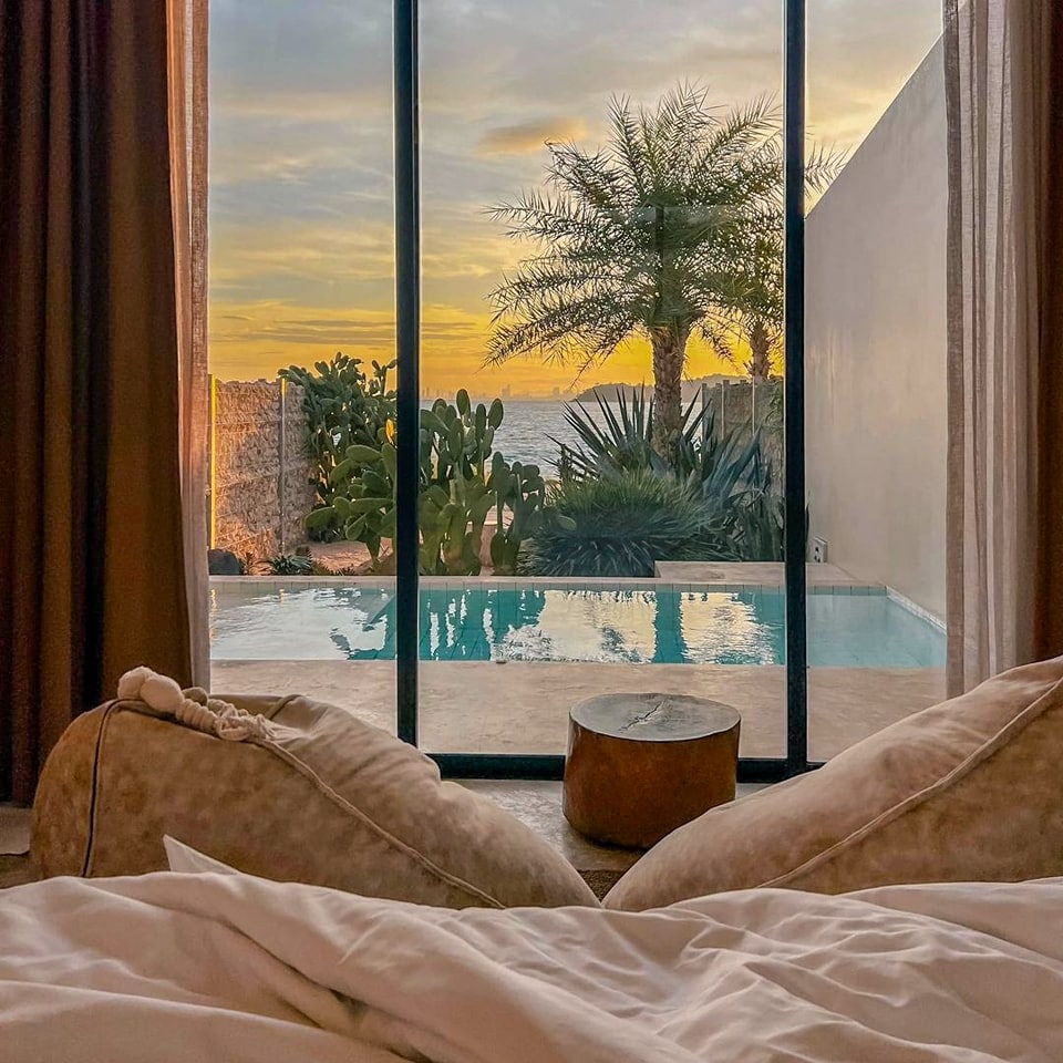 The view from the interior features a serene scene of a private pool framed by palm and cactus plants. Warm sunset hues illuminate the sky beyond large glass doors, while a cozy bed with soft pillows is partially visible in the foreground.