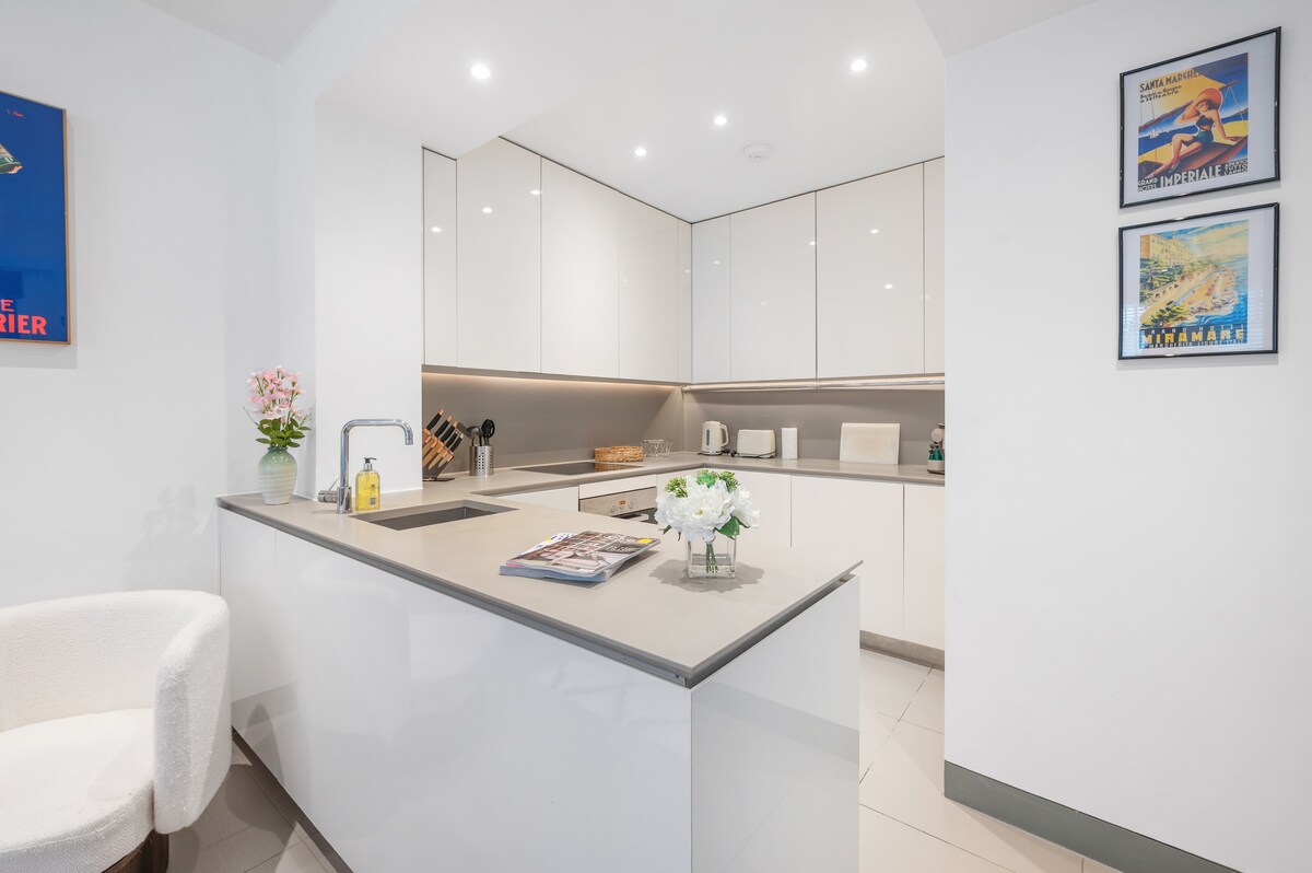 A modern kitchen area is showcased, featuring sleek white cabinetry and countertops. Essential appliances like a kettle and microwave are arranged neatly, with a small floral arrangement adding a touch of decor. Bright lighting enhances the clean aesthetic of the space.