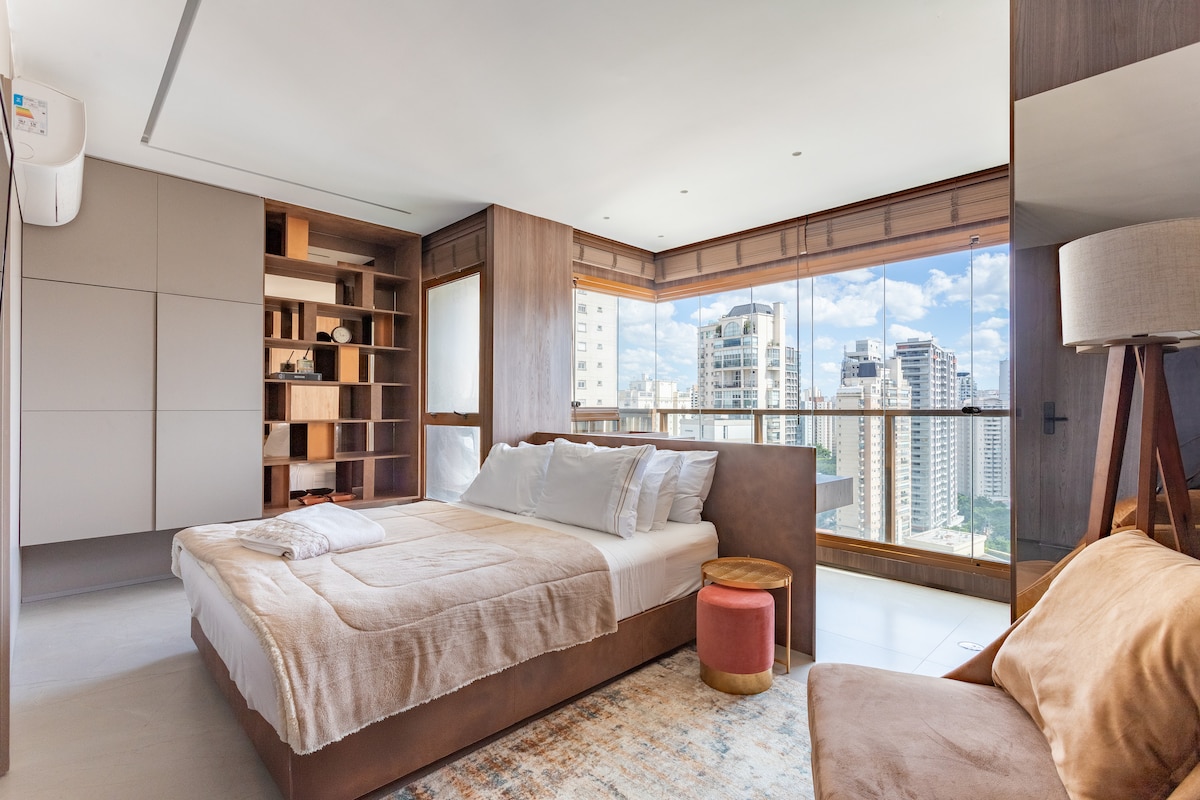 A spacious bedroom features a large queen bed with soft bedding. A wooden accent wall adds warmth, while a built-in bookshelf provides storage. Natural light floods the room through a large glass balcony, offering panoramic views of the cityscape. A small side table is placed next to the bed.
