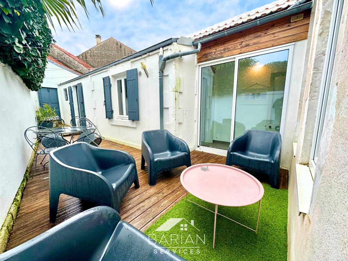 A private outdoor terrace features a smooth wooden deck and artificial grass. Four comfortable armchairs surround a round pink table, creating a cozy outdoor seating area. The walls are painted white, and light streams in from large glass doors, enhancing the space's openness.