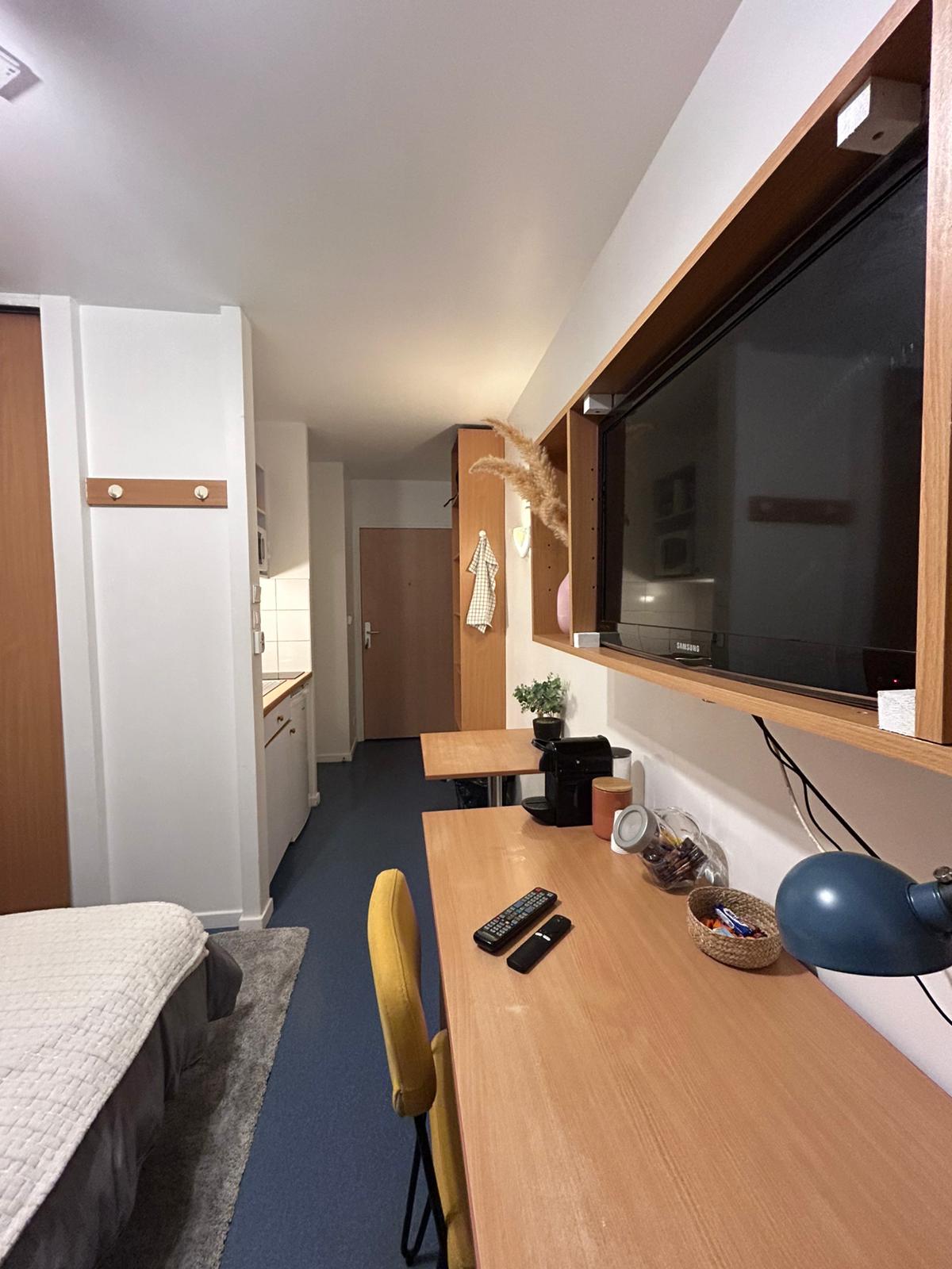 The image captures a cozy studio featuring a spacious bed along one side and a desk with a bright yellow chair on the opposite side. A television is mounted on the wall above the desk, and various decorative items are arranged neatly. The kitchenette and bathroom doors are visible in the background.
