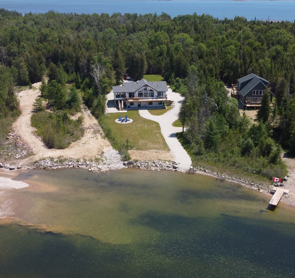 An aerial view of the cottage situated on the shores of Lake Huron, surrounded by green trees and a sandy beach. The spacious driveway leads to the entrance, while a small path traces along the water's edge, framing the tranquil inlet.