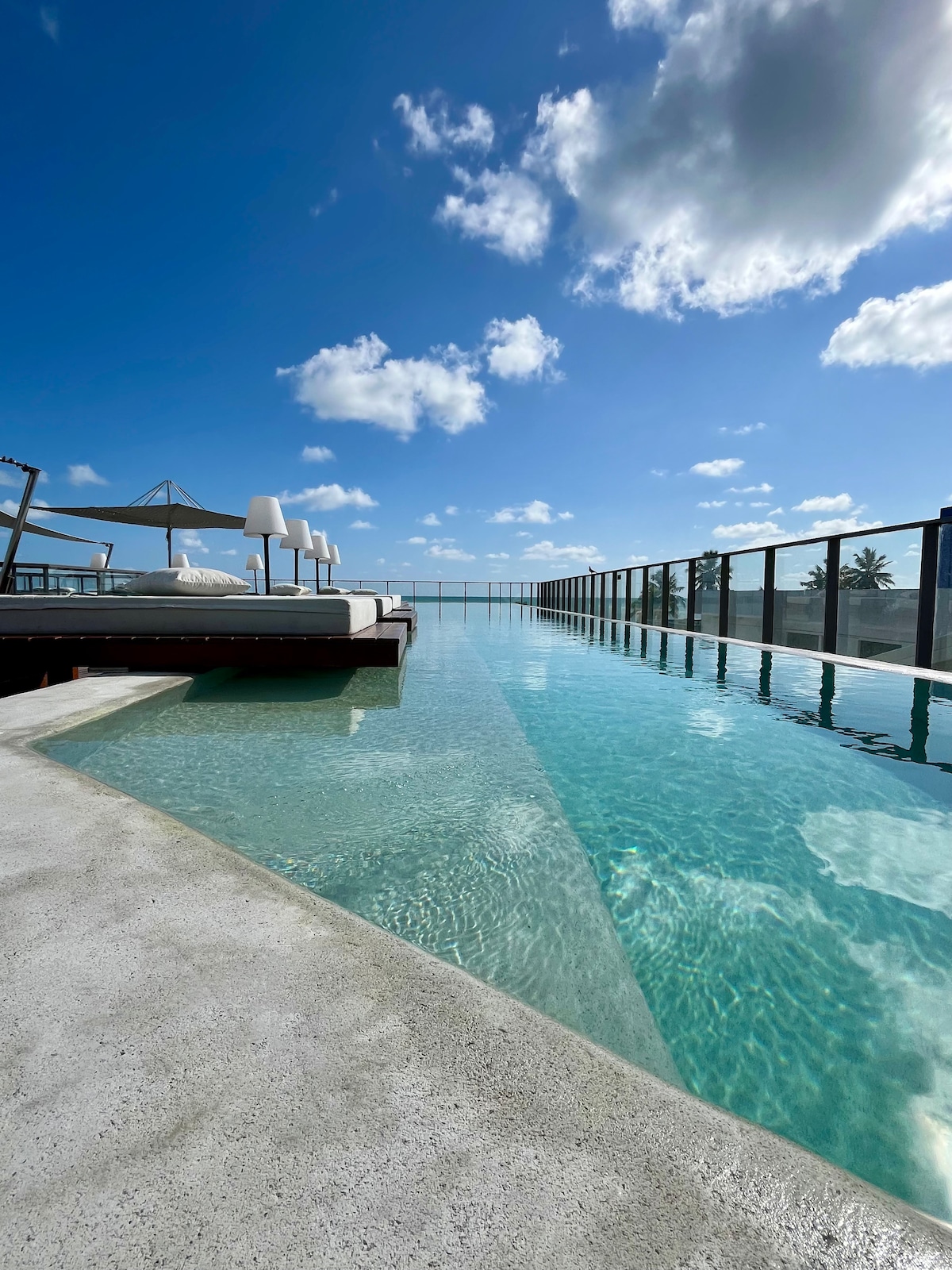 A panoramic view of an infinity pool extends towards the horizon, bordered by a sleek glass railing. Sunbeds with umbrellas offer a relaxing space nearby, under a clear blue sky dotted with fluffy white clouds.