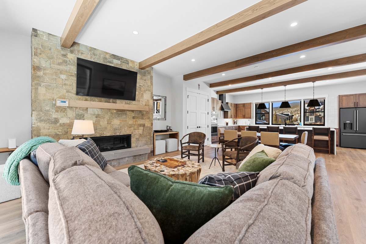 The open living area features a spacious sectional sofa with decorative cushions, set against a stone accent wall containing a flat-screen TV. Natural light enters through large windows, revealing a glimpse of the modern kitchen and dining area with stylish bar stools.