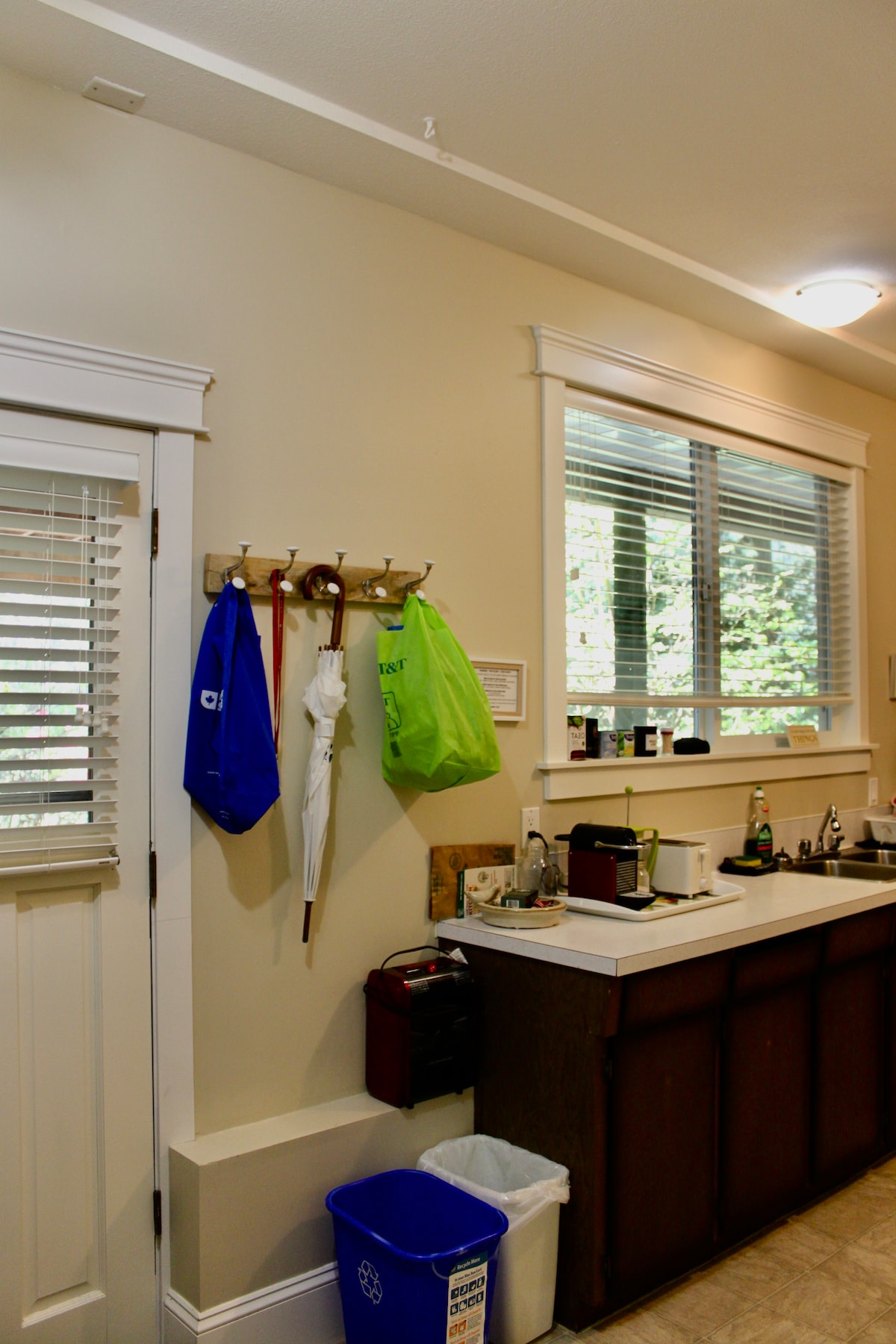 A well-organized kitchen area is depicted, featuring a countertop with various kitchen appliances and utensils. Coats and bags hang on hooks by the door, while windows allow natural light to illuminate the space. A trash bin is positioned near the cabinetry.