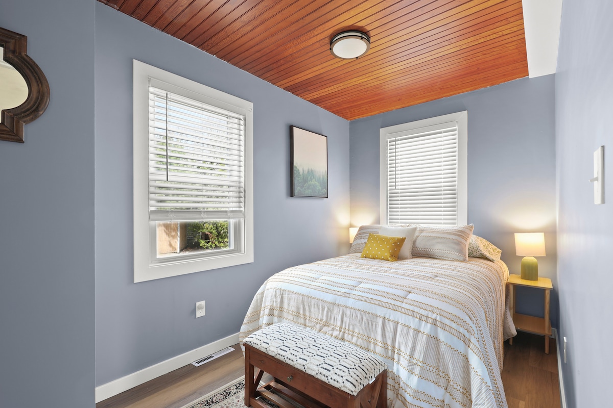 A serene bedroom features a spacious bed adorned with a textured blanket. Natural light filters through two windows with white blinds. Warm wood paneling complements the soft blue walls. A small bedside table and a decorative bench provide additional storage and comfort.