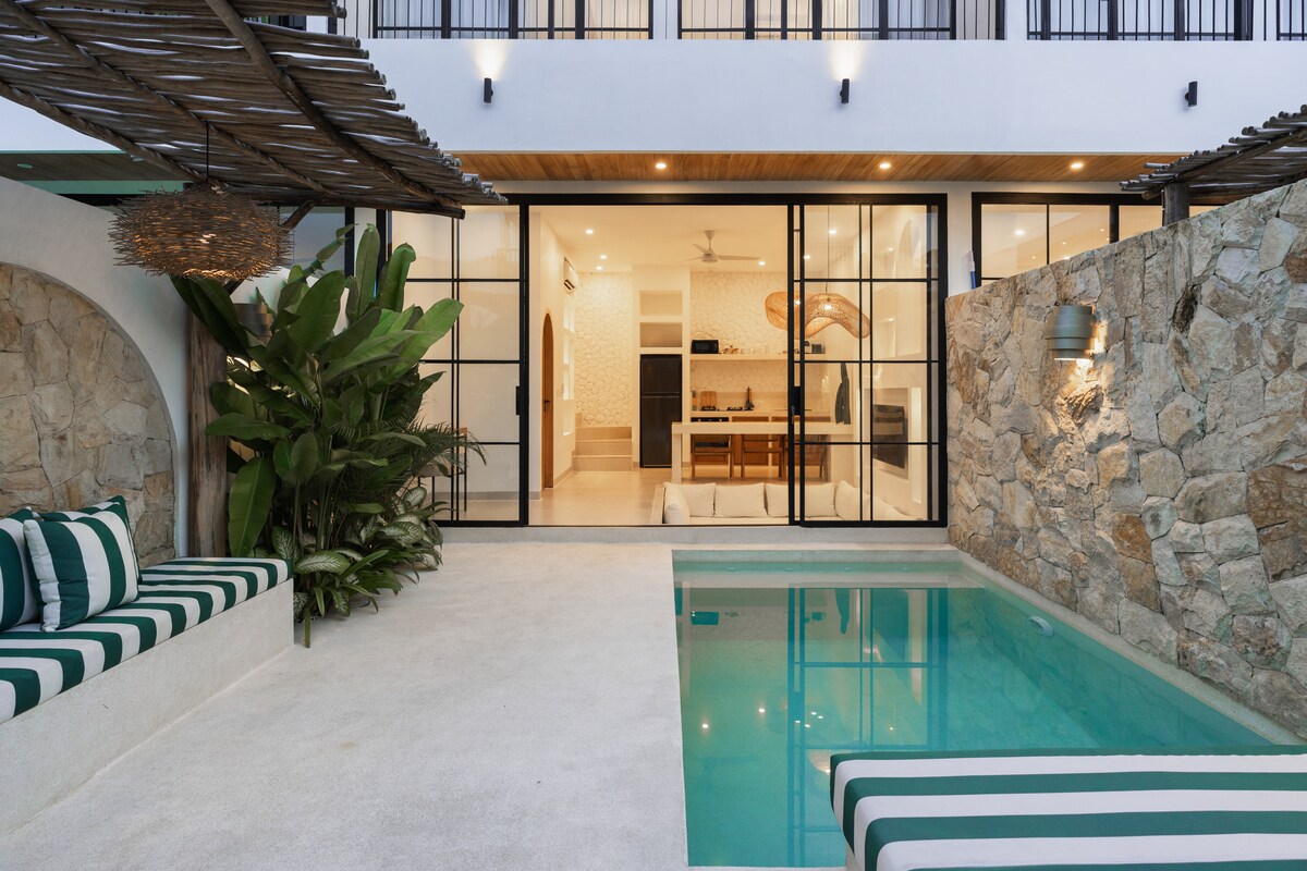 An outdoor area highlights a private pool bordered by smooth stone walls. A sunken couch with green and white stripes sits nearby, surrounded by lush tropical plants. Large glass doors open to reveal the open-plan living space, seamlessly connecting the outdoors with the interior.