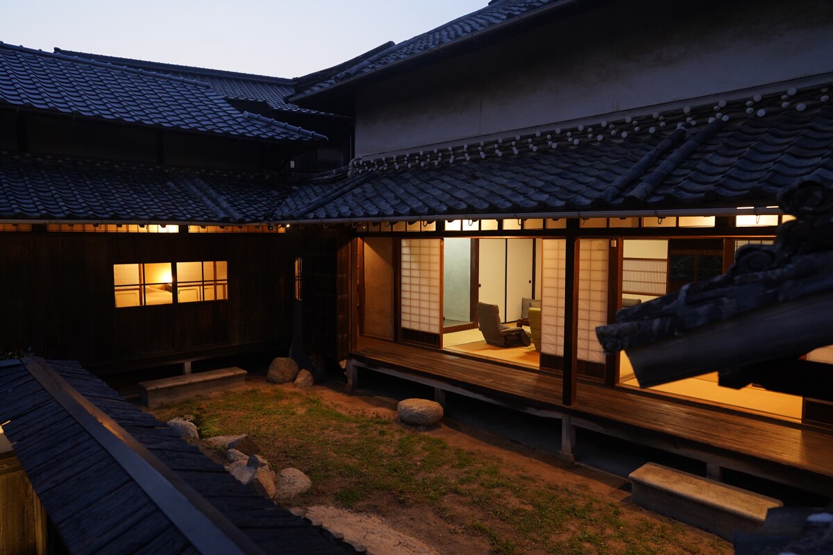An evening view captures the traditional architecture of the house, showcasing wooden beams and a lush green garden. Soft lighting casts a warm glow from the windows, illuminating the interior spaces while the muted colors of dusk envelop the scene.