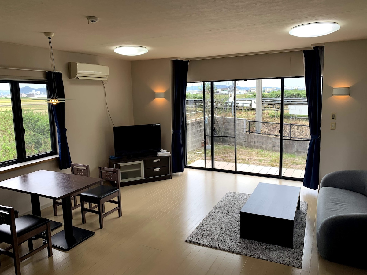 A bright living area is presented with a sliding glass door that offers views of the outdoor space. A dark wood dining table with four chairs is positioned beside a cozy gray sofa. A television sits on a low cabinet, illuminated by recessed lighting.