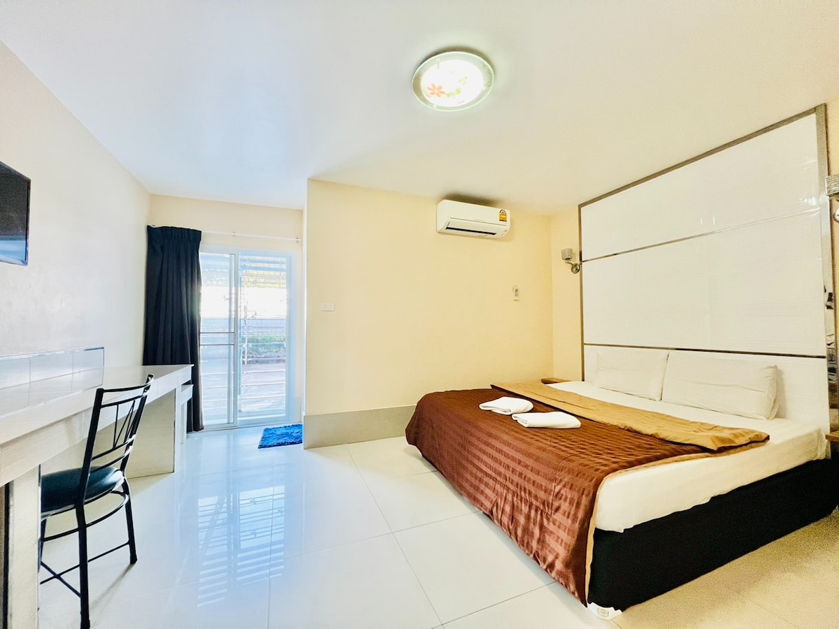 A bright room features a spacious bed with crisp bedding and warm-toned throw. A simple desk with a chair is positioned against the wall, while large glass doors provide access to an outdoor area. Air conditioning and a flat-screen TV are also visible.