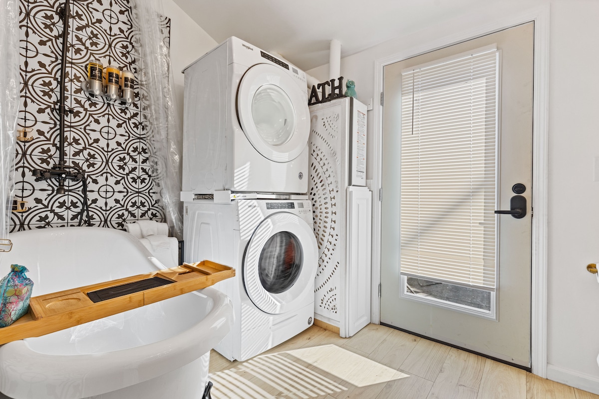 A bright laundry area features a stacked washer and dryer, providing convenience for guests. A stylish freestanding bathtub is positioned next to the washer, and a wooden tray rests atop the tub. A door with blinds allows natural light to enter the space.