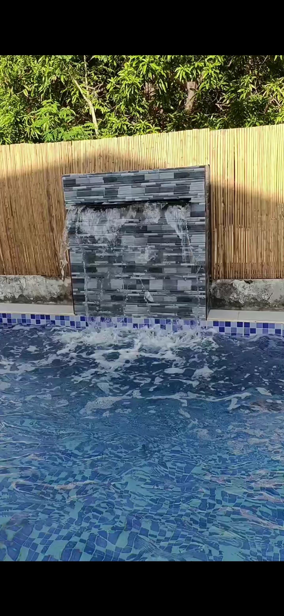A water feature is seen cascading into a clear blue pool, creating gentle ripples on the surface. The backdrop features a textured gray stone wall, flanked by bamboo, which provides a natural setting for relaxation.