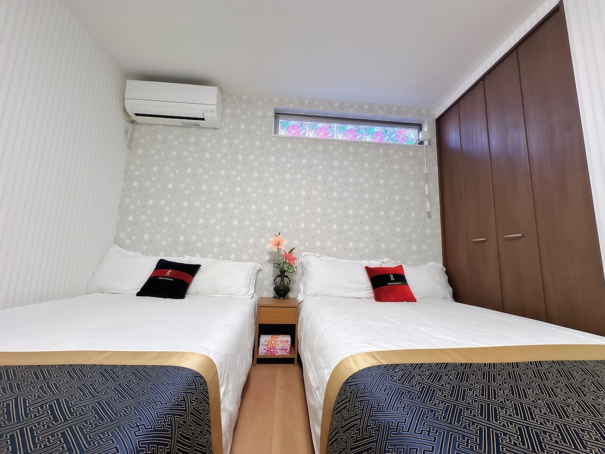 The room contains two spacious beds with decorative black and gold bedspreads. A small bedside table holds a flower arrangement. Light enters through a window adorned with floral curtains, while a wardrobe is positioned against the wall, contributing to the room's organized appearance.