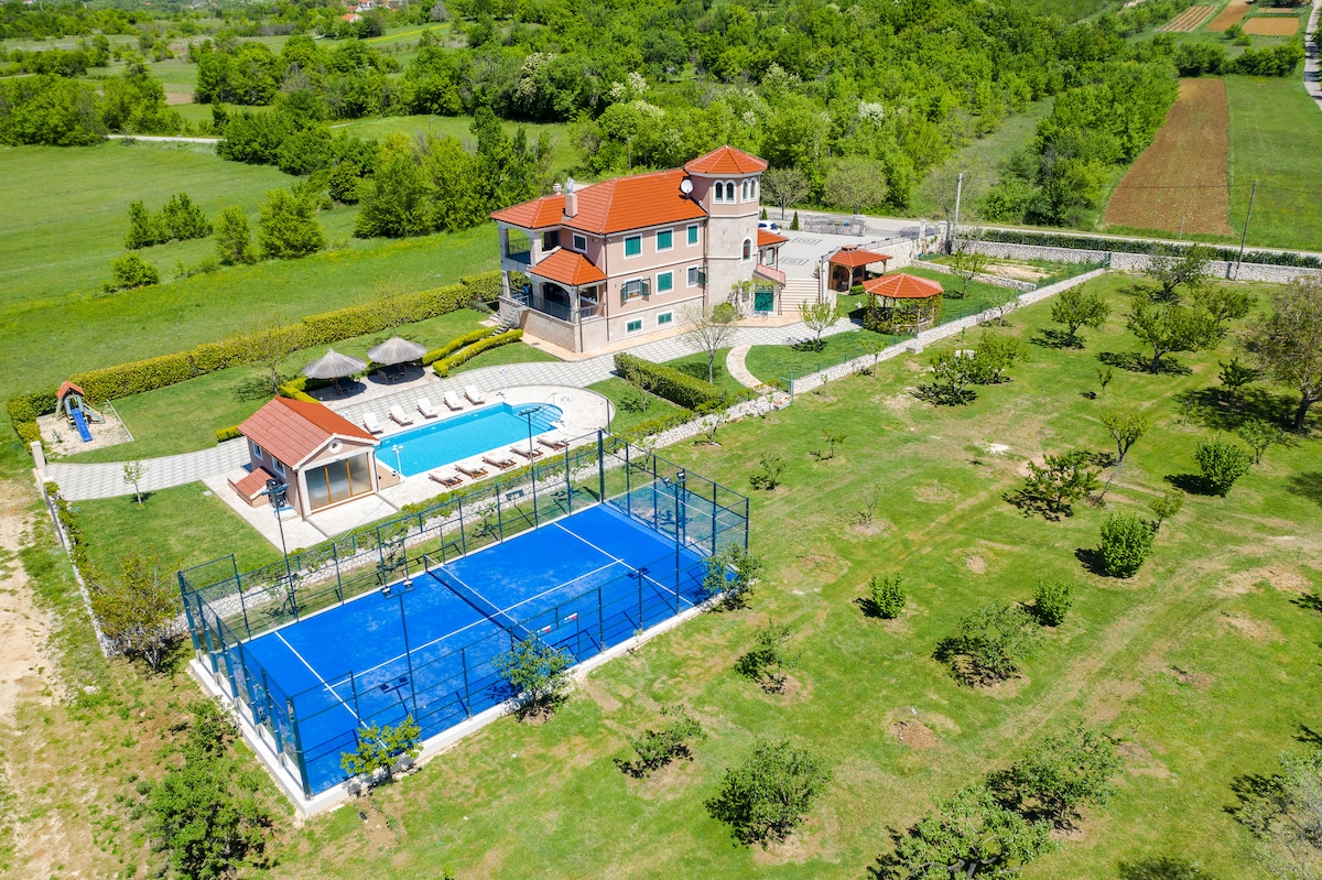 An aerial view of a spacious villa set in the lush greenery of the Dalmatian countryside, featuring a heated pool surrounded by sun loungers. A blue padel court is visible alongside the well-maintained lawn and gardens. The property includes several outdoor seating areas.