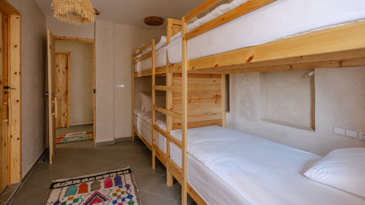 A shared sleeping area features wooden bunk beds with clean linens, providing ample accommodation. A colorful woven rug adds a warm touch to the gray tiled floor. Two doors lead to other spaces, creating a sense of connectivity in the dorm.