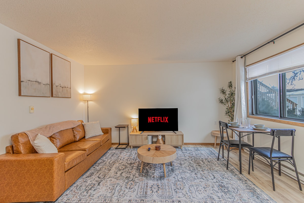 A spacious living area features a large, comfortable sofa and a 50" Smart TV displaying Netflix. A round coffee table sits in the center, with a dining table for two nearby. Soft lighting from a floor lamp creates a warm ambiance.