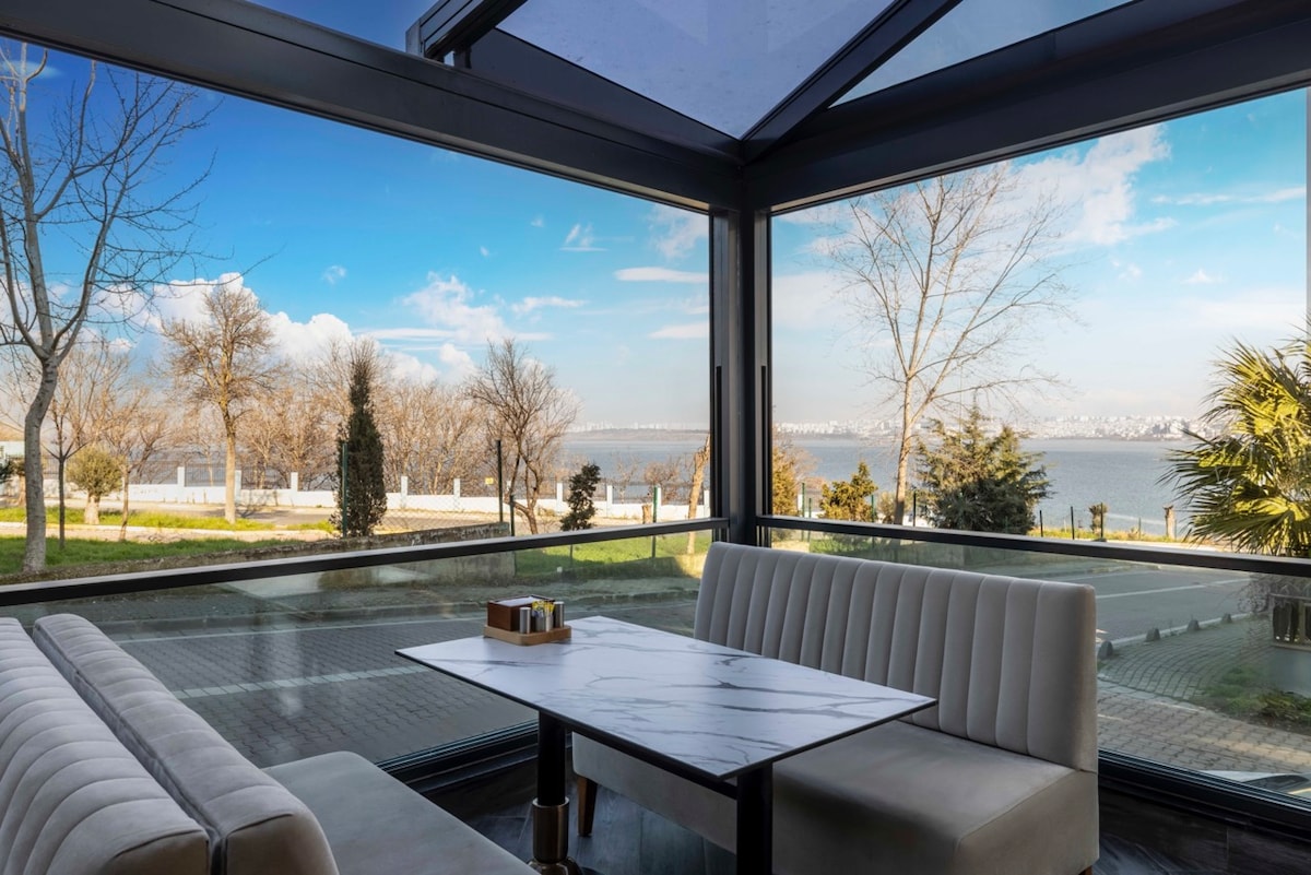 A contemporary seating area is highlighted by large glass windows offering expansive views of the lake and surrounding greenery. A marble table rests between plush benches, creating a comfortable space to enjoy the serene scenery outside.