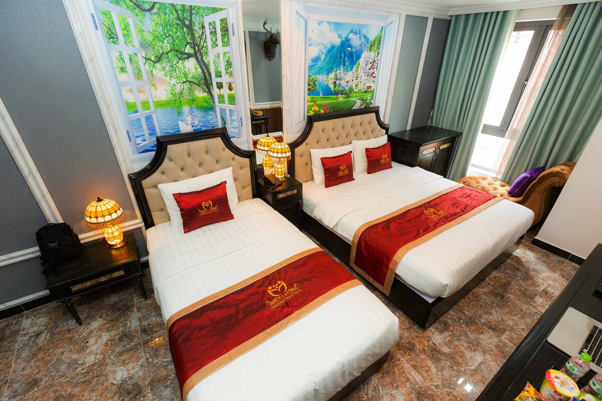 A well-lit guest room features two spacious beds dressed in white linens and decorative red accents. Large wall art depicting serene landscapes enhances the ambiance, while elegant bedside lamps provide soft illumination. Dark curtains frame the windows, and a cozy seating area is visible in the corner.