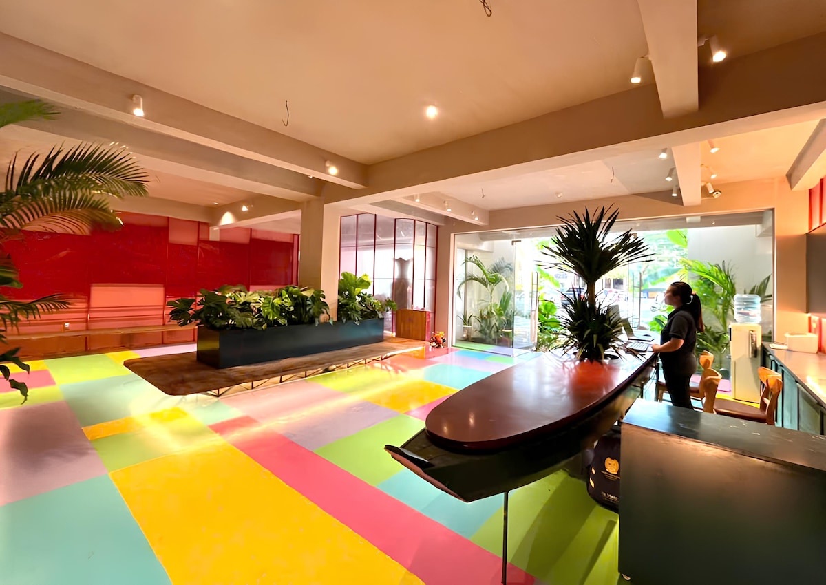An open communal area features a vibrant, multicolored floor with spacious seating and a collection of lush green plants. Natural light streams in through large windows, creating a bright and inviting environment that encourages social interaction.