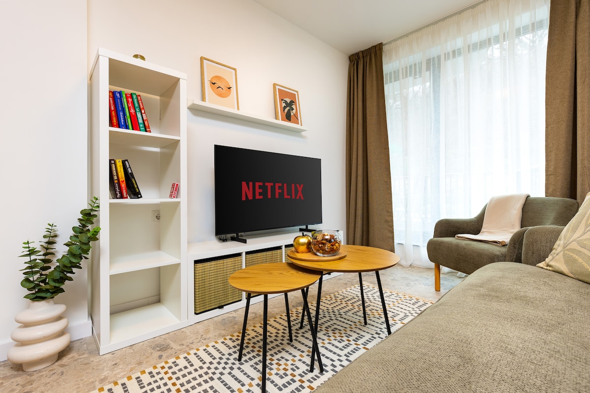 A cozy living area features a soft, gray sofa paired with two round wooden coffee tables. A flat-screen television displays the Netflix logo on the wall opposite. Light filters through sheer curtains, enhancing the warm tones of decorative elements and bookshelves.