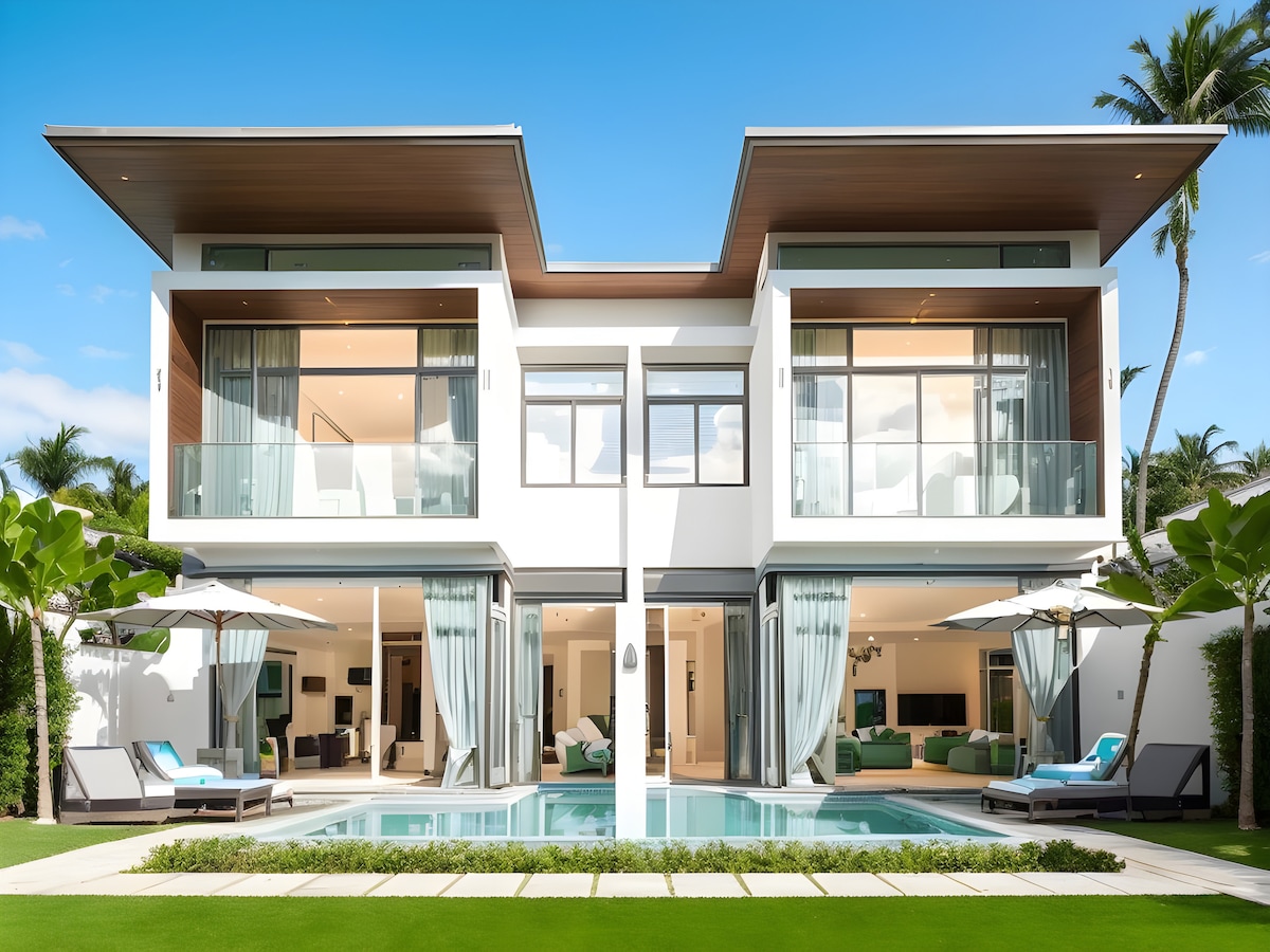 A modern two-story villa is featured, showcasing a bright façade with expansive glass doors flanked by balconies. Lush tropical greenery surrounds the private pool, which reflects the clear blue sky. Outdoor seating areas with umbrellas invite relaxation by the water.