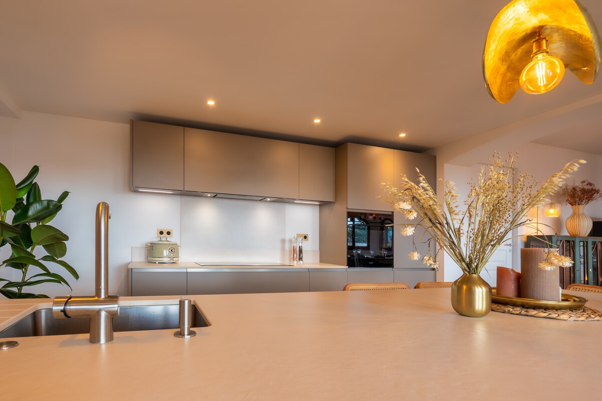 A modern kitchen is showcased, featuring sleek cabinetry and a spacious countertop. A decorative vase with dried flowers complements the scene. Task lighting is provided by a unique pendant light, while a green plant adds a touch of nature to the setting.