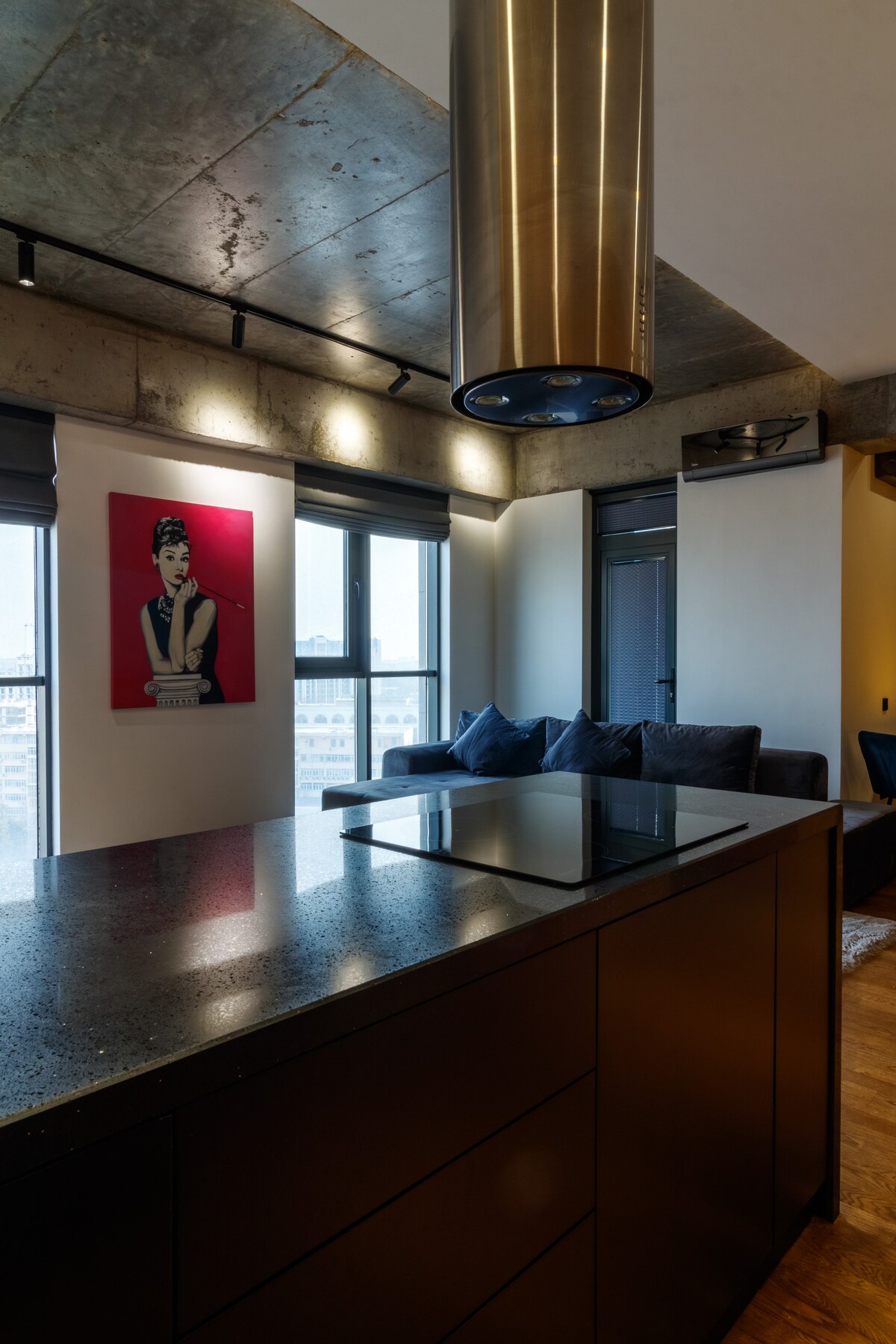 The image showcases a contemporary kitchen featuring a dark countertop and an embedded stovetop. A spacious living area is visible in the background, highlighted by large windows and a stylish artwork on the wall. Soft lighting enhances the modern atmosphere of the space.