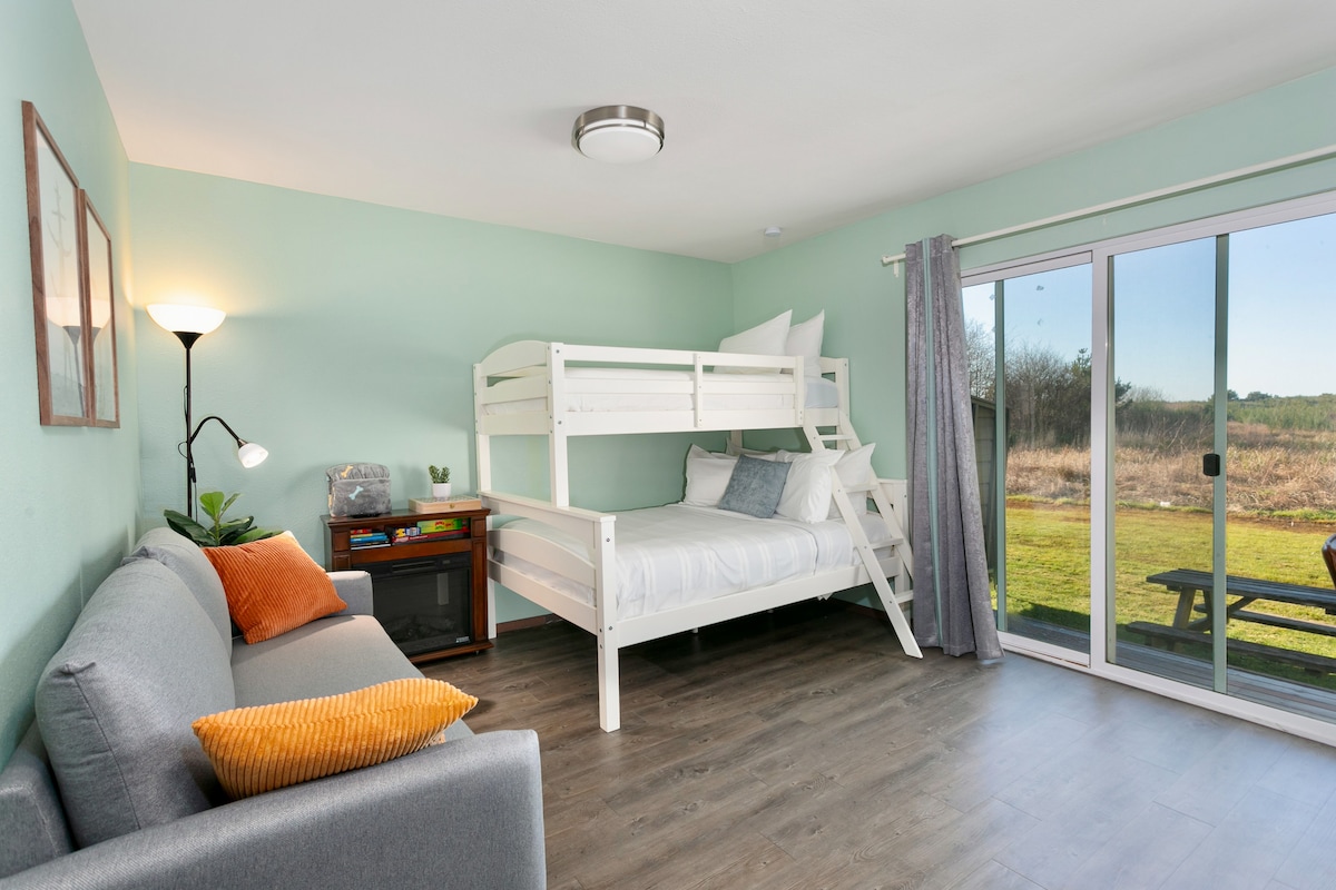 The bright main room features a cozy seating area with a gray sofa and orange accent pillow. A white bunk bed is visible, with a spacious bed beneath. Full patio doors open to a grassy landscape, allowing natural light to fill the room.