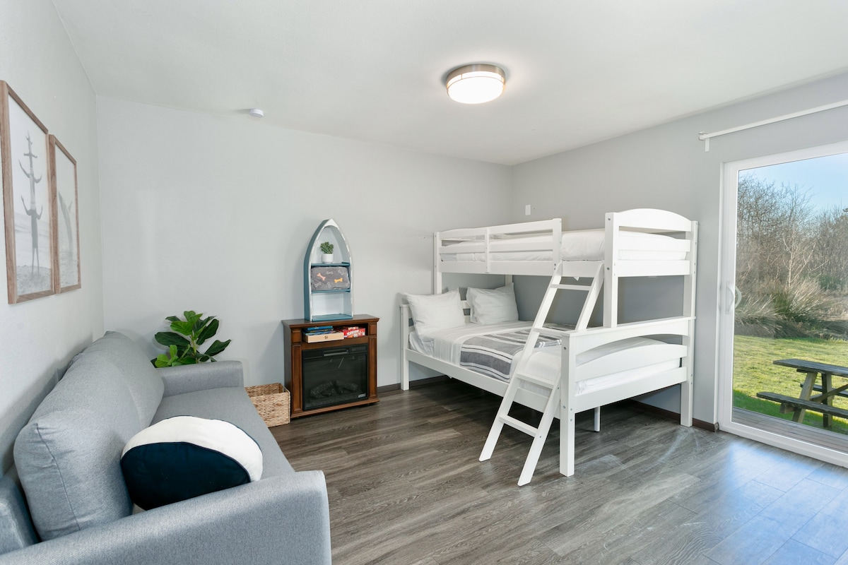 A comfortable living space features a light-colored sofa and a set of white bunk beds. A cozy electric fireplace is situated next to a decorative shelf. Full patio doors open to a view of the outdoors, allowing natural light to fill the room.