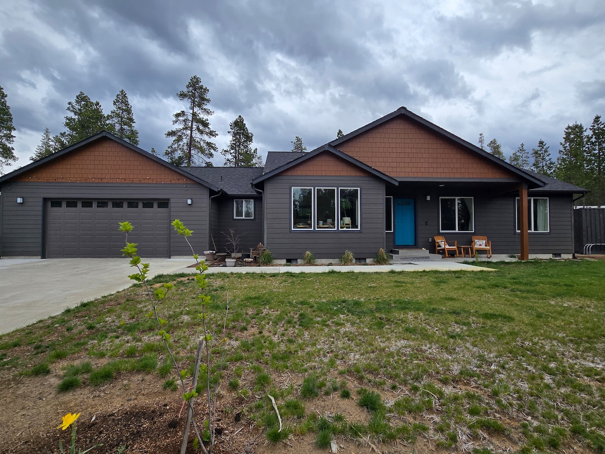 The exterior view of the country home is presented, showcasing a modern design with a blend of dark and light siding. A spacious driveway leads to a two-car garage. Large windows allow natural light to fill the interior, while a seating area is visible on the front porch.