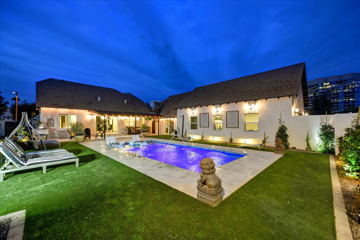A well-maintained outdoor area features a vibrant blue pool surrounded by plush green grass. Lounge chairs are positioned near the water, while soft lighting highlights the exterior walls of the home. The twilight sky enhances the serene ambiance of this inviting space.
