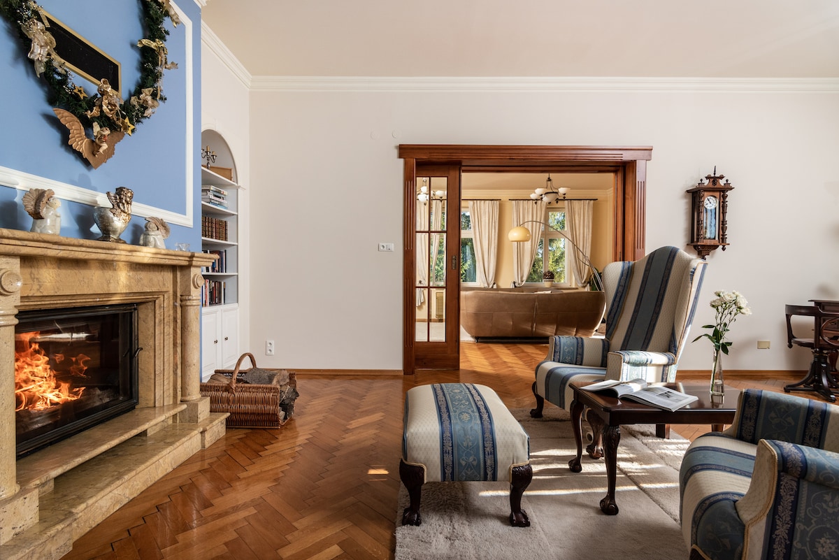 A warm living area features elegant seating with striped upholstery and a cozy stone fireplace. Natural light filters through glass doors, revealing a glimpse of a separate room adorned with soft furnishings. Decorative elements include a wall-mounted clock and a simple floral display.