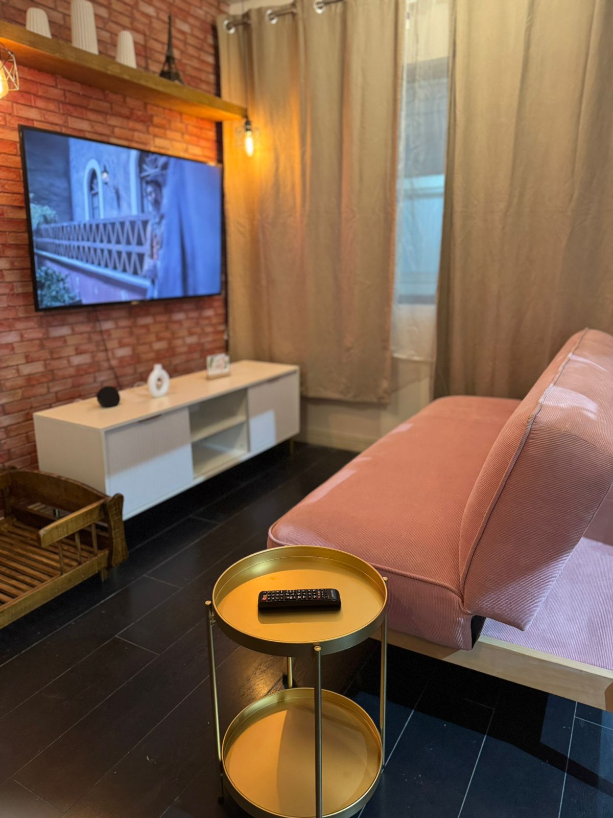 A living area features a soft pink sofa facing a wall-mounted television. A small gold table is positioned nearby next to a wooden tray, creating a functional space. The floor is dark, and the room is adorned with light brown curtains that complement the exposed brick wall.