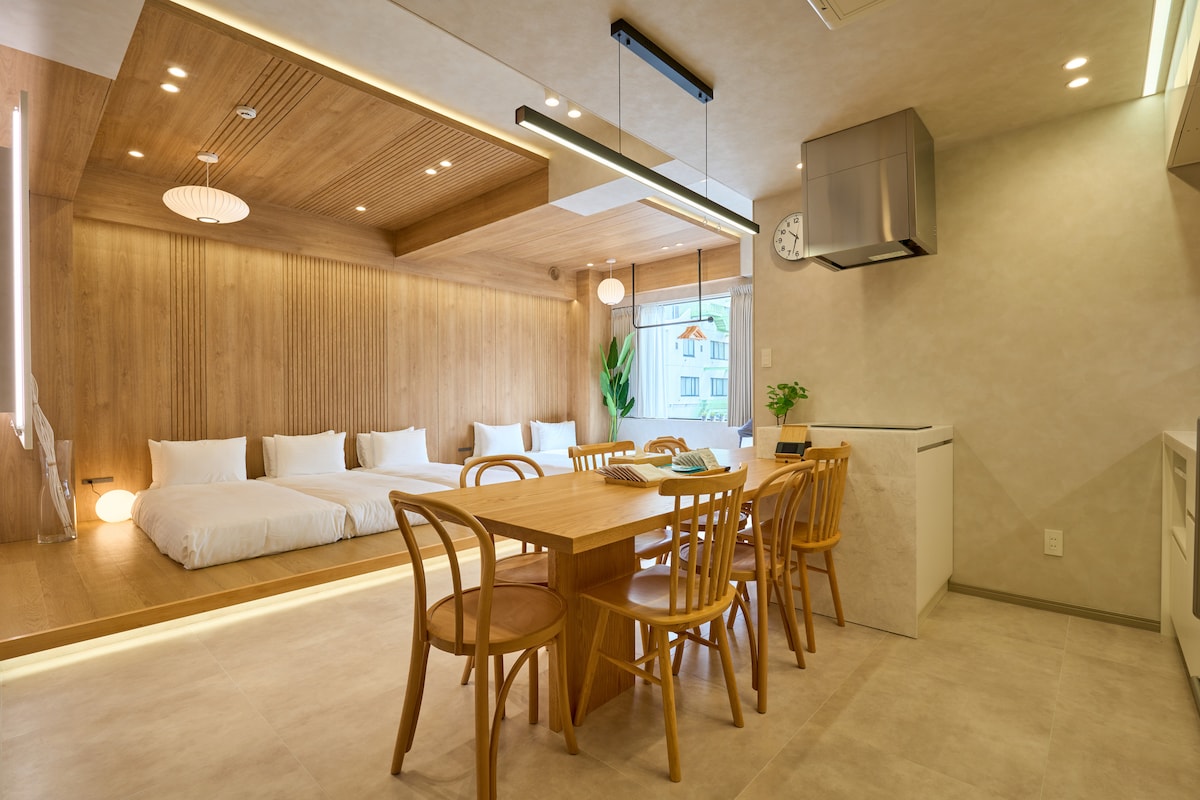 The image features a modern living space with natural wood accents. A dining table with wooden chairs occupies the center, while a spacious sleeping area with low-profile bedding is visible in the background. Soft lighting creates a warm ambiance, complemented by greenery from a plant near the window.
