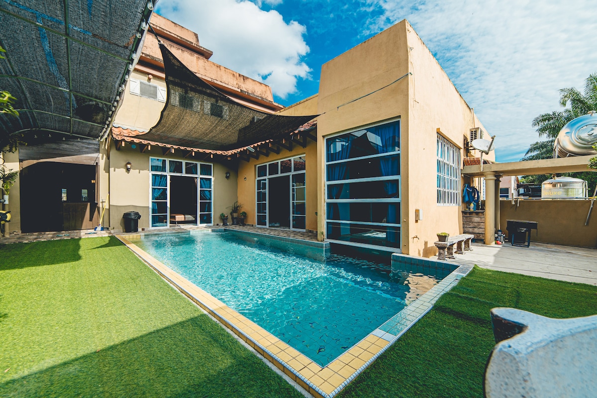 An expansive private swimming pool is surrounded by well-maintained grass and outdoor seating. Large glass doors provide views into the villa, where natural light illuminates the space. A covered area offers protection from the sun, contributing to a relaxed atmosphere.