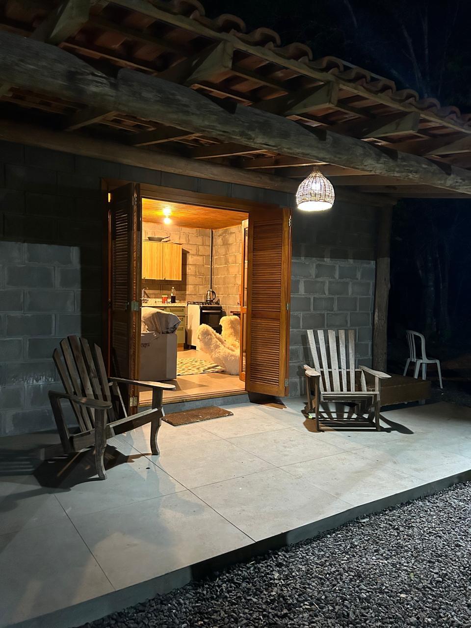 The entrance of a cozy kitchen can be seen through open wooden doors, inviting guests inside. Two wooden chairs are positioned on a stone patio, illuminated by warm overhead lighting. A dog is visible in the kitchen, enhancing the homey atmosphere.