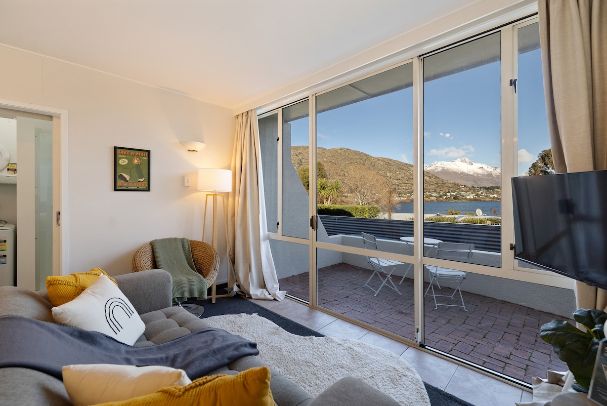 A cozy living area includes a comfortable sofa adorned with decorative cushions. Large sliding glass doors provide panoramic views of Lake Wakatipu and the surrounding mountains. Soft natural light illuminates the space, highlighting the contemporary decor and inviting atmosphere.