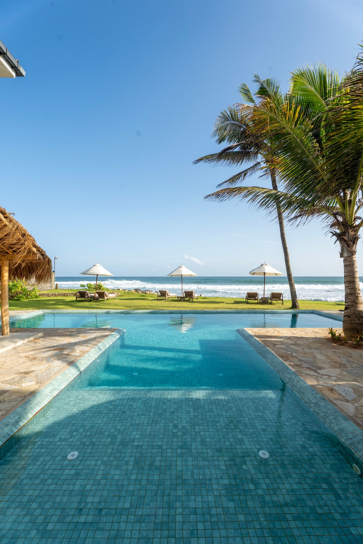 A clear view of a tranquil swimming pool bordered by palm trees. Beach umbrellas are positioned beside the pool, overlooking the ocean waves. The serene environment is enhanced by the bright blue sky and lush green grass, creating a relaxing outdoor space.