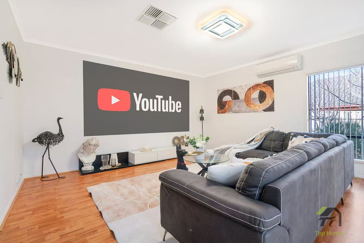 A modern living area features a large wall-mounted projector displaying the YouTube logo. A sectional sofa is placed in front, complemented by decorative accents such as sculptures and artwork. Natural light enters through vertical blinds, illuminating the warm wood flooring.