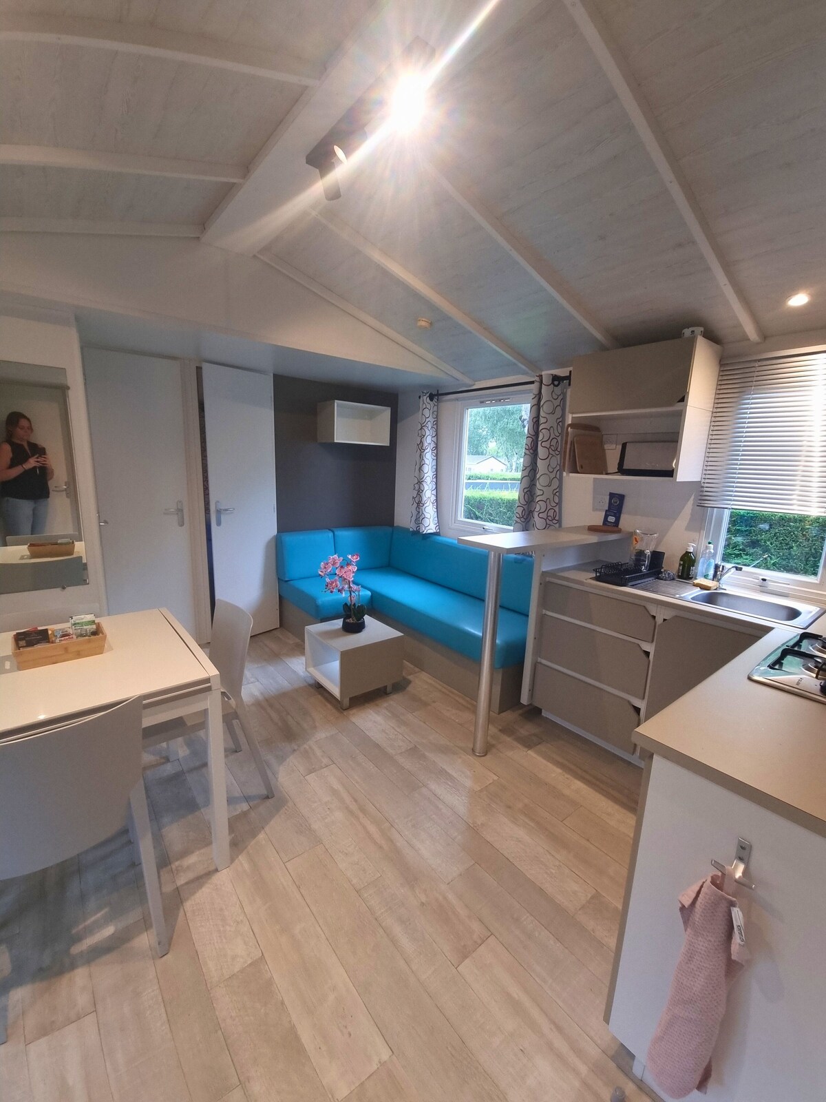 A bright and spacious interior of the mobil-home is shown, featuring a comfortable teal sofa that invites relaxation. The open layout includes a dining table and a fully equipped kitchen. Natural light filters through the window, enhancing the modern decor and airy atmosphere.