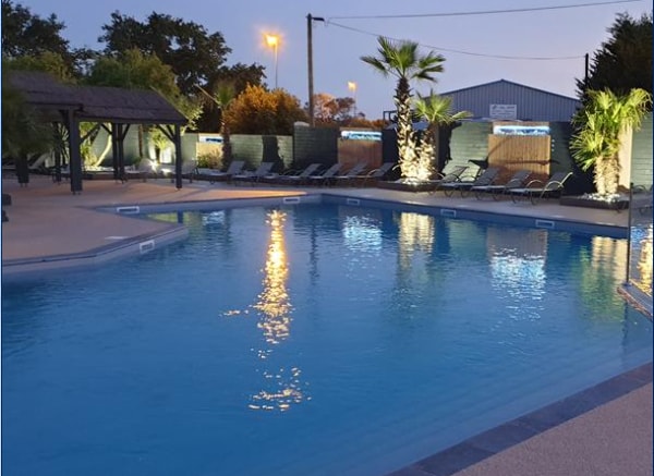 An inviting swimming pool is surrounded by palm trees and well-lit seating areas. The water is calm and reflects the warm lights from the nearby structures, creating a serene environment for relaxation. Lounge chairs are positioned around the pool for comfortable seating.