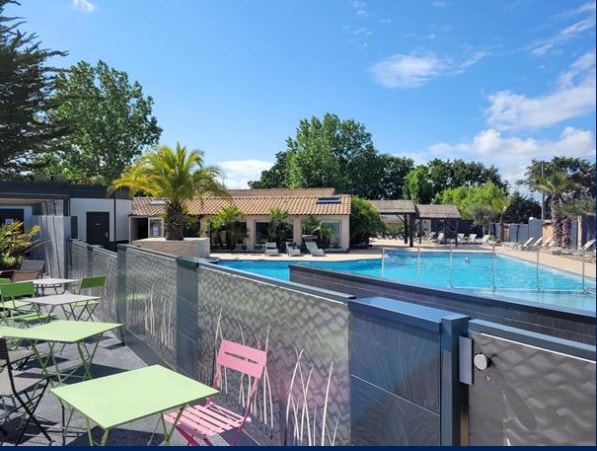 A swimming pool is visible, surrounded by sun loungers and tropical greenery. Colorful tables and chairs in shades of green and pink are set nearby, offering inviting seating options. The pool area is framed by trees and a charming structure that adds to the relaxing atmosphere.