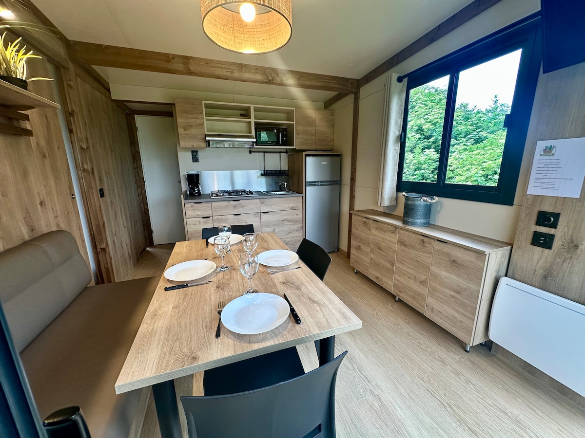 The kitchen area features modern appliances and ample cabinetry, creating a functional space for meal preparation. A dining table is set with plates and glasses, complemented by natural light streaming through large windows, enhancing the cozy atmosphere of the tiny house.