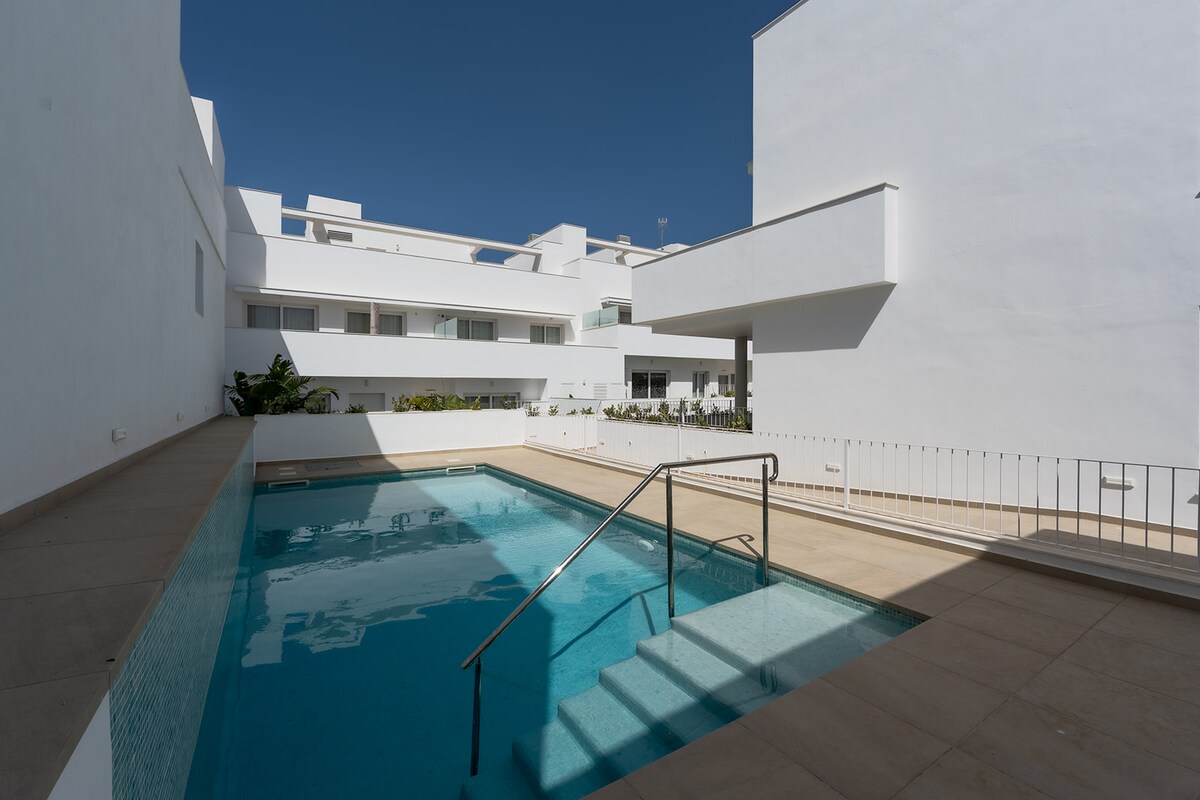 A community pool is present within a bright, open area, surrounded by white buildings. Clear water is visible, with steps leading into the pool. Sunlight reflects off the surface, creating a serene setting for relaxation outdoors.